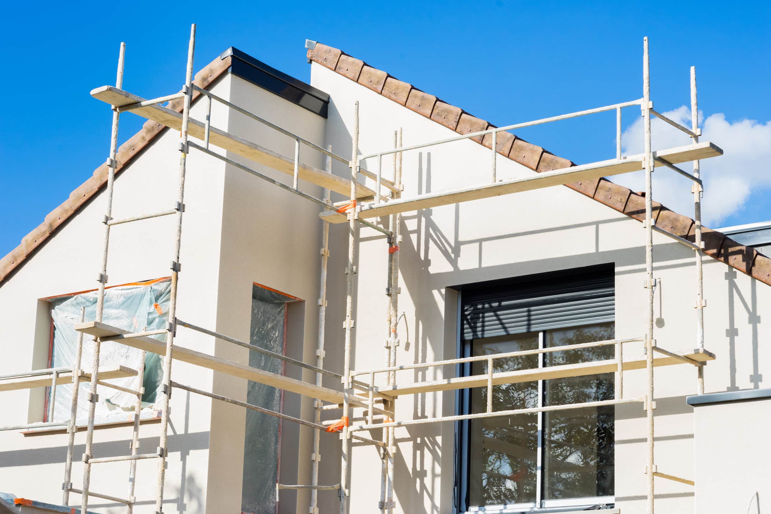 Chantier de construction : échafaudage devant une maison neuve crépie, fenêtres protégées sous plastique, sous ciel bleu.