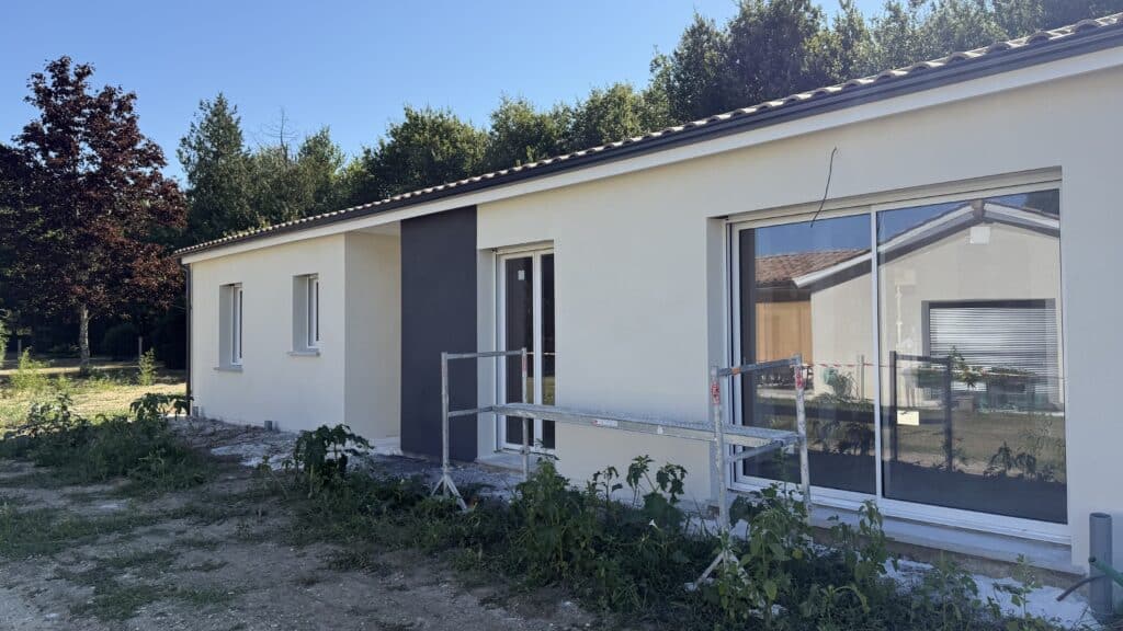 Maison neuve contemporaine : chantier et façade Maison neuve avec enduit clair et accent gris anthracite, fenêtres modernes et échafaudage devant, chantier bordé d'arbres et ciel bleu.
