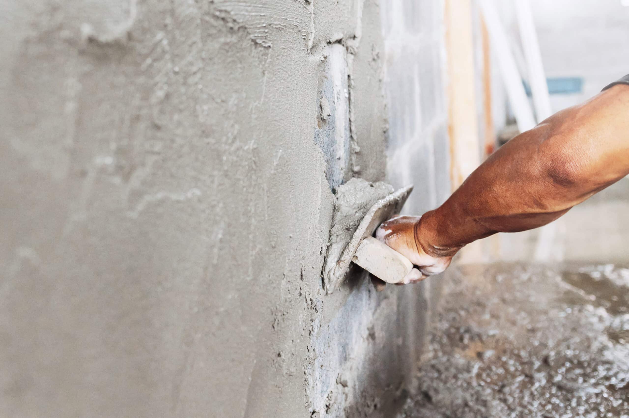 Application de mortier : Rénovation murale artisanale Main foncée appliquant du mortier sur un mur en parpaings avec une truelle, travaux de ragréage en cours.